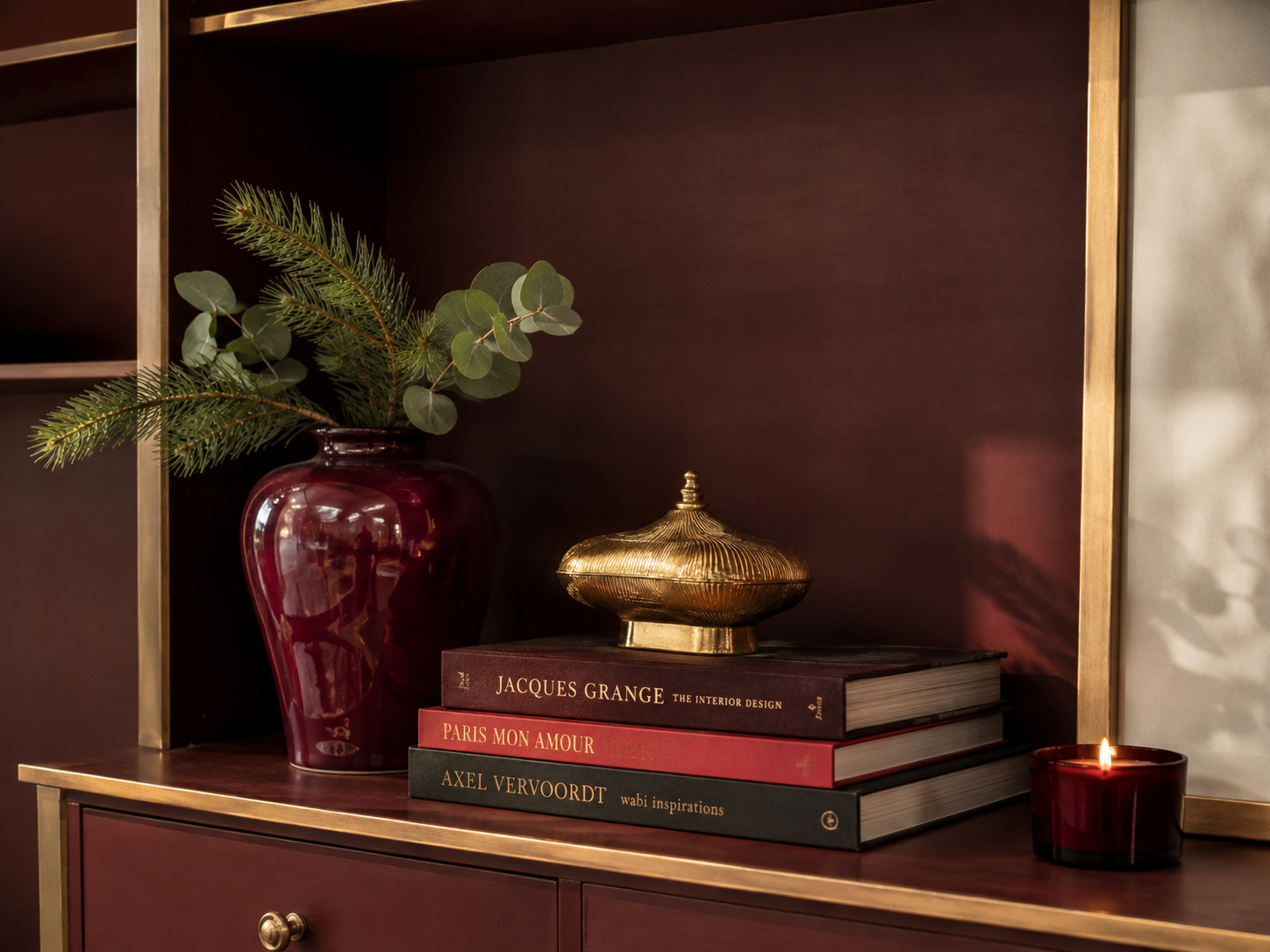 Composition bordeaux et or sur meuble avec vase profond, livres, objet en laiton et petite bougie.