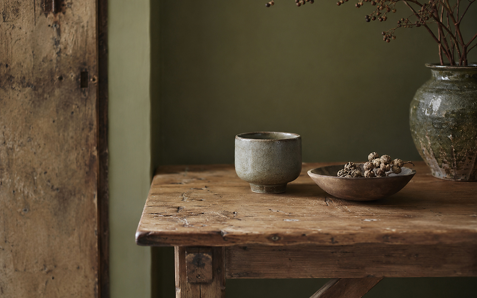 Table en bois brut devant un mur vert olive avec céramique artisanale et branches séchées.